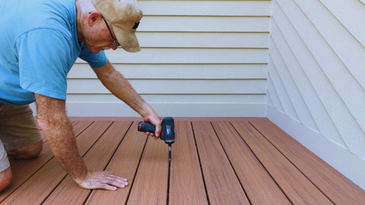 A man, identified as a decking contractor, operates a drill while installing a wooden deck in a backyard setting.